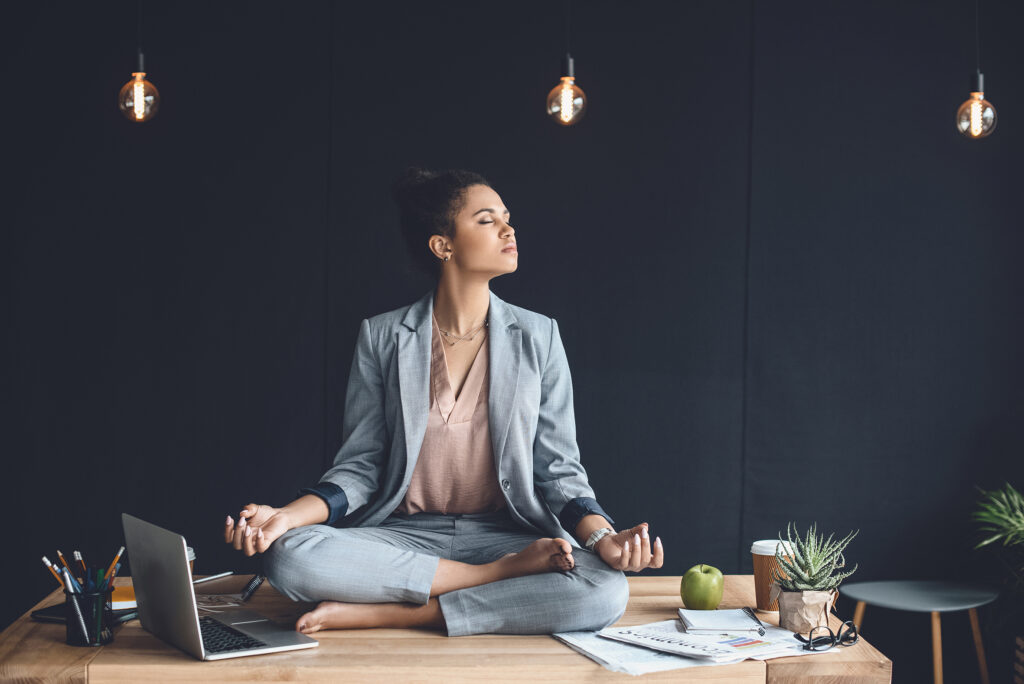 african american businesswoman sitting in lotus pose on table while meditating in office