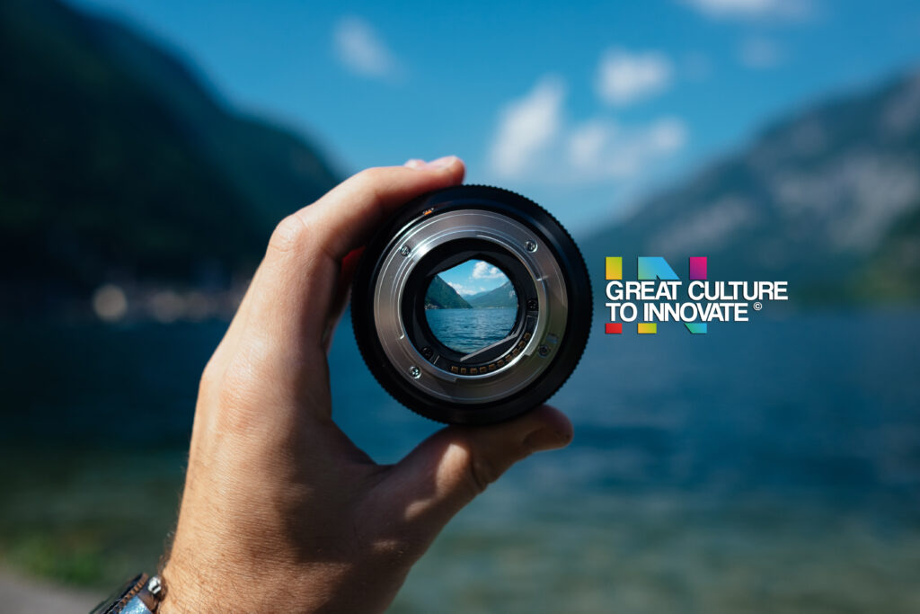 man holding camera lense infront of a beautiful sea between mountains