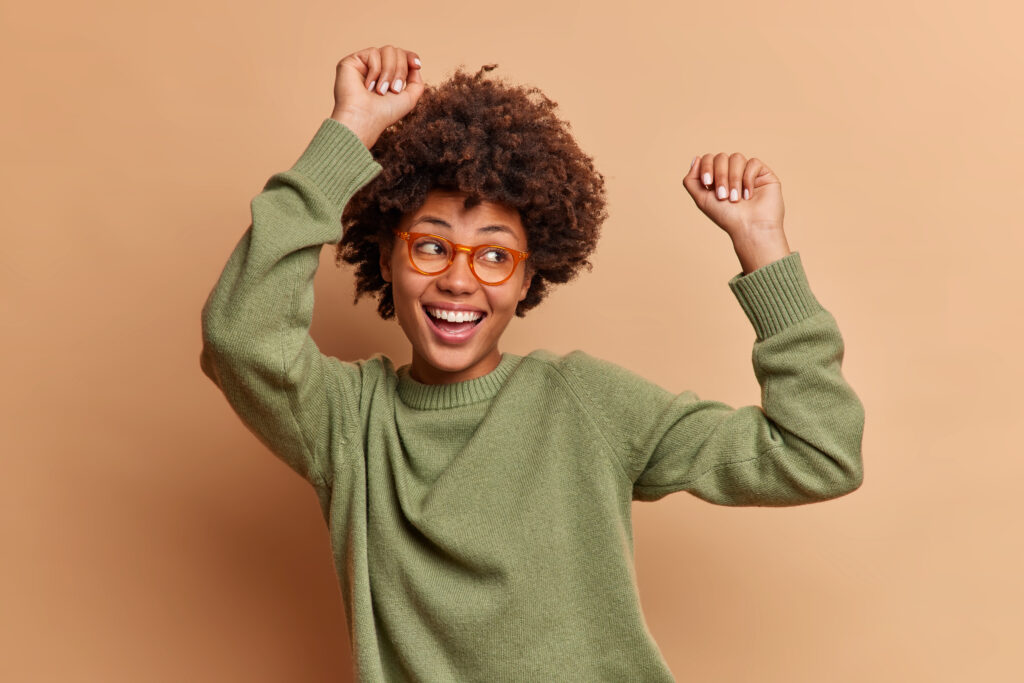 Magnificent glad dark skinned woman dances with arms raised laughs happily being full of energy enjoys favorite music looks aside dressed casually isolated over brown background. Happy dancing female