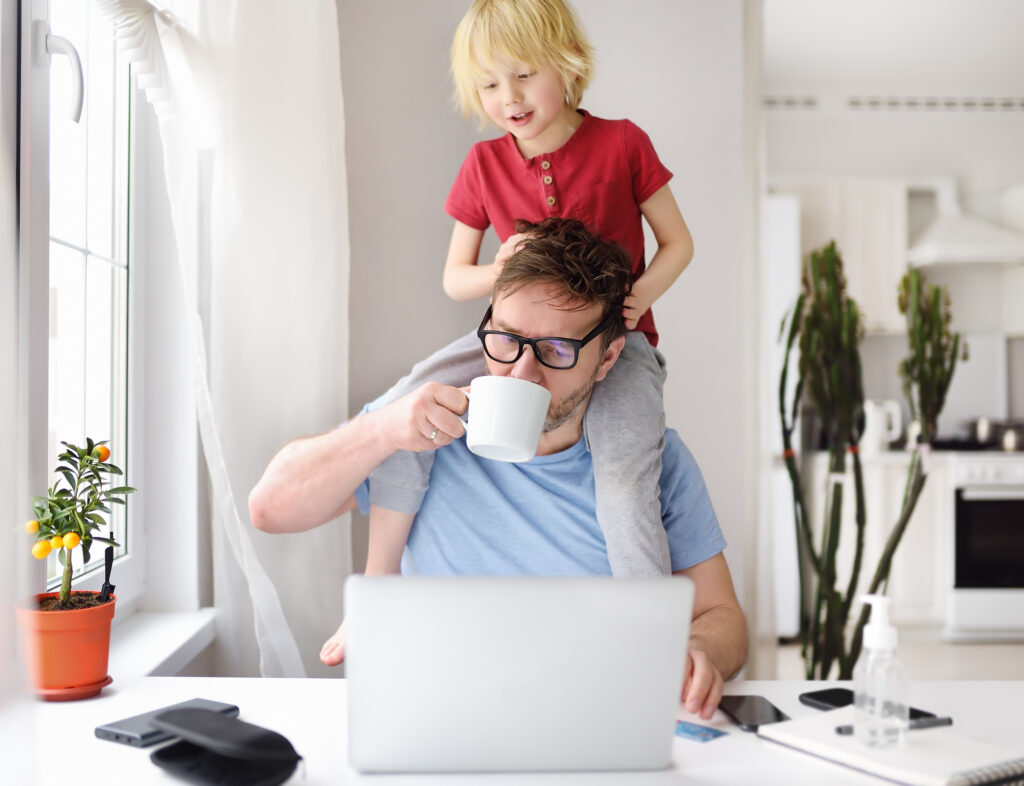 man working on a laptop while having his child on his shoulders