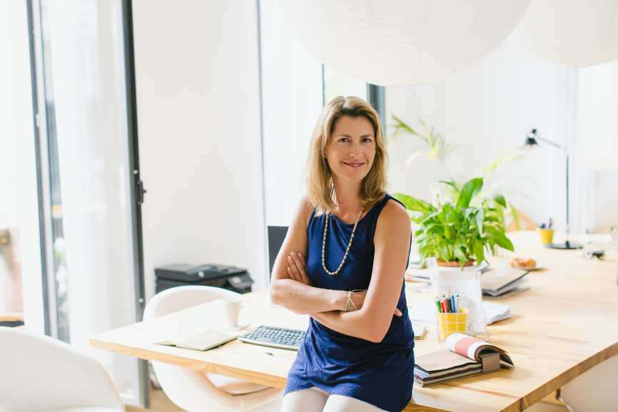 Frau mit blauem Kleid und blonden Haaren lehnt sich an einen Bürotisch