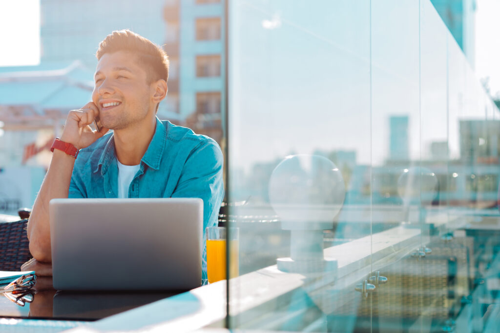 Joyful young man wearing casual attire smiling while having a pleasant conversation with his friend and sitting at his laptop at an urban cafe.